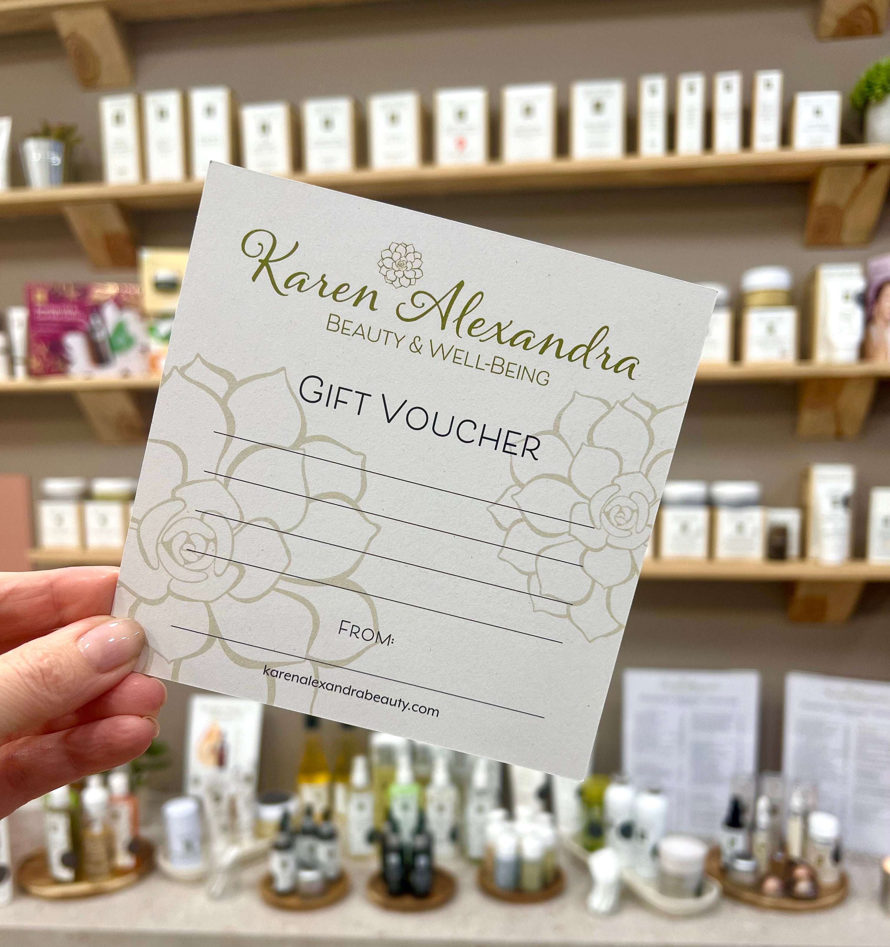 A physical gift voucher from Karen Alexandra Beauty and Well-Being Tonbridge with a floral pattern held in front of a shelf of beauty products.
