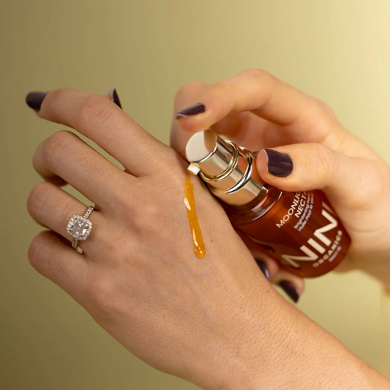 Hand applying NINI Organics moonlight nectar brown cosmetic product from a bottle on a beige background