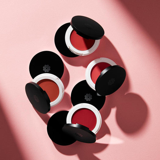 Multiple compact pans of Lily Lolo Lip and Cheek Cream in various shades, displayed on a pink background.