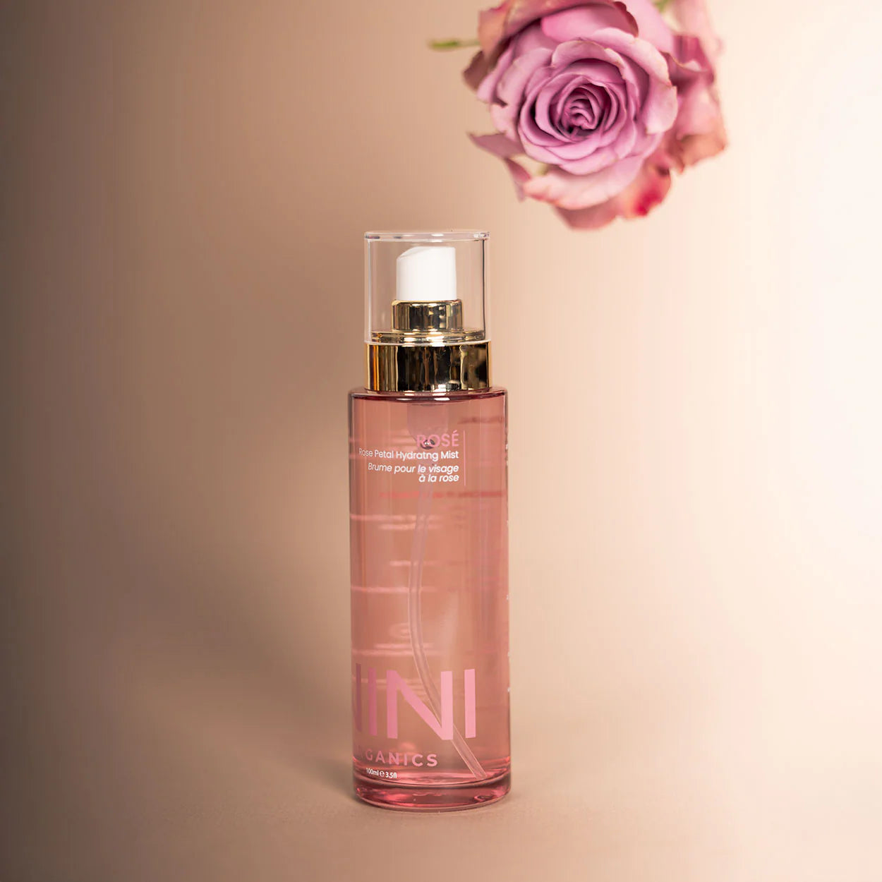 NINI Organics Rose Water Face Mist Skincare bottle with gold cap and pink liquid on a beige background with a pink rose.