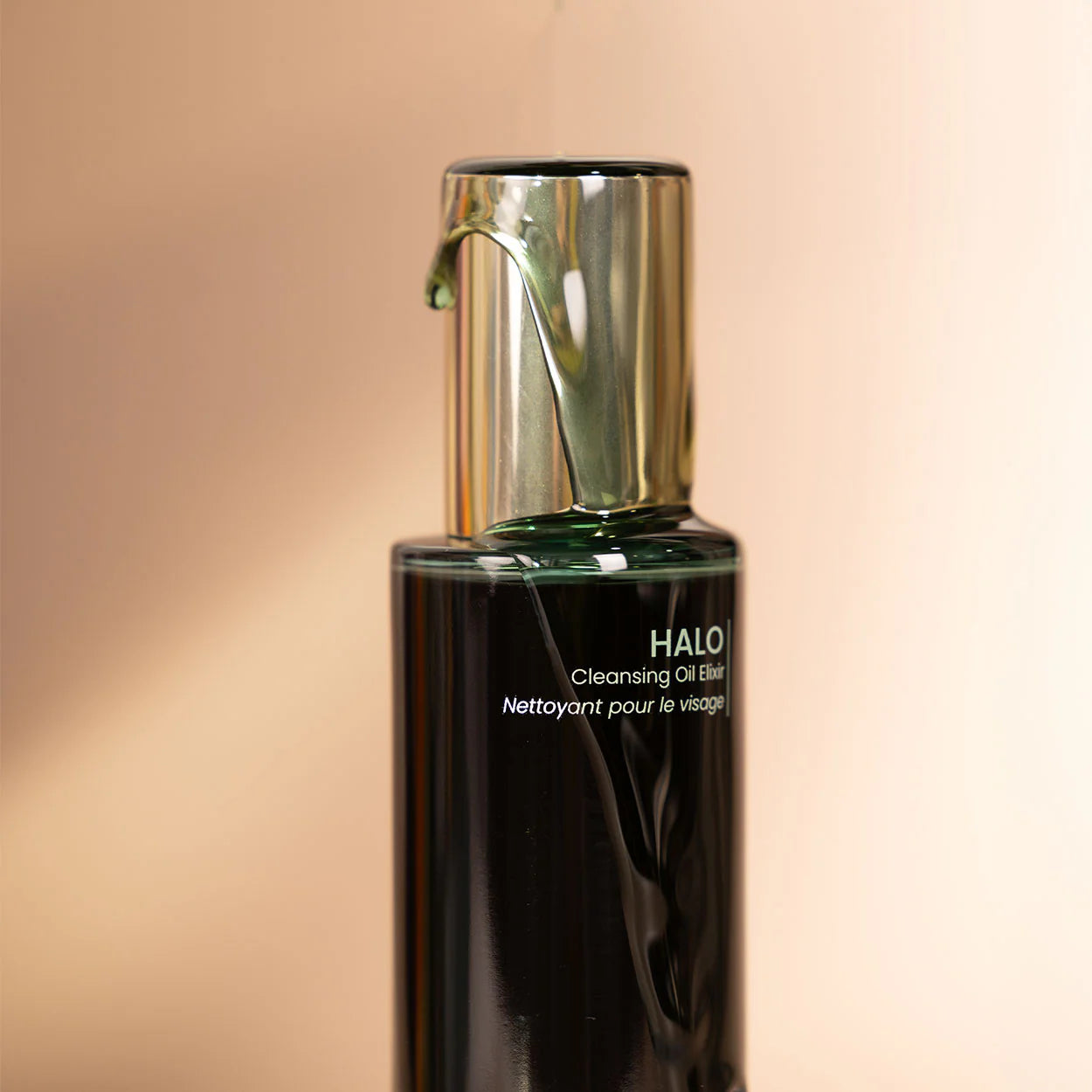 Black bottle of Halo Cleansing Oil with a metallic cap on a beige background with oil being dispensed